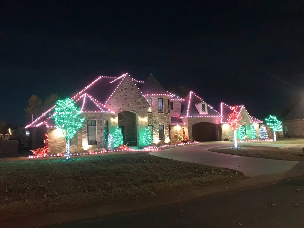 A large two-storey stone home outlined in red and white bulbs, with trees wrapped in green and red and lights edging the driveway.